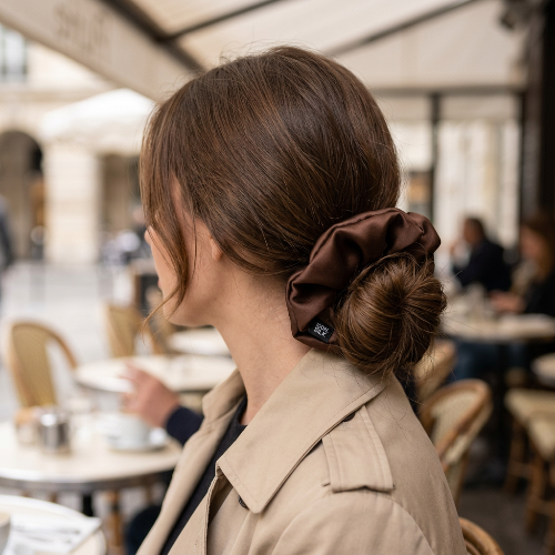 Chouchou en cheveux naturels cafe porte autour d un chignon bas par une femme brune en manteau beige sur une terrasse