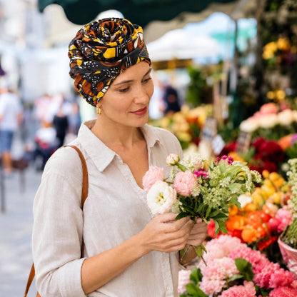 Turban pour femme a motifs jaunes noirs et marron porte avec un bouquet de fleurs en main