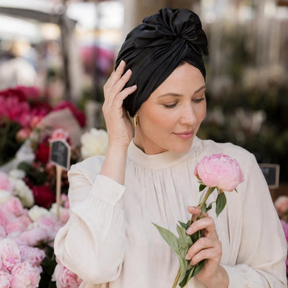 Turban pour femme noir avec noeud sur le devant porte par une femme tenant une fleur rose